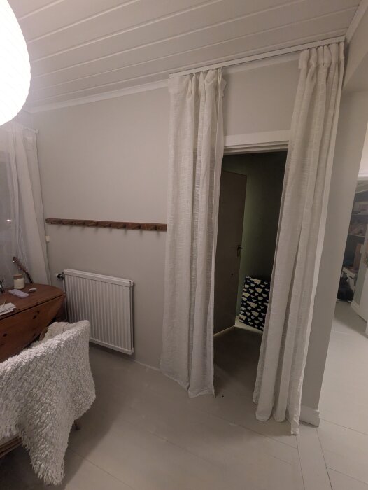 Rum med vita väggar och tak, vita gardiner vid en dörröppning, en vit radiator och ett träskrivbord med ullfilt över stolen.