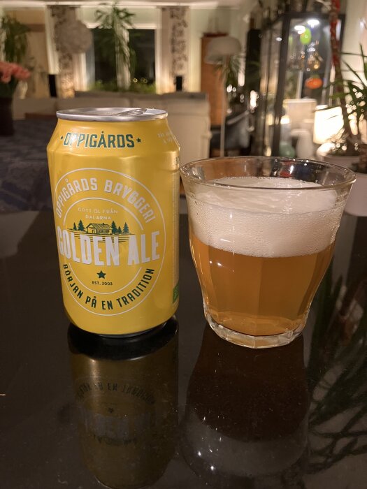 En burk Oppigårds Bryggeri Golden Ale och ett glas med öl står på ett bord. Bakgrunden visar ett vardagsrum med växter och lampor.