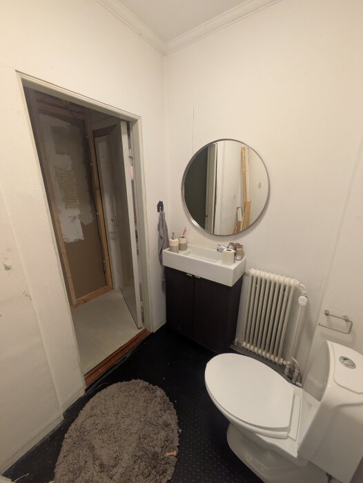 Badrumsinteriör med toalett, handfat, spegel och oavslutad dörröppning till höger. Svart golv, vitmålade väggar och synligt element.