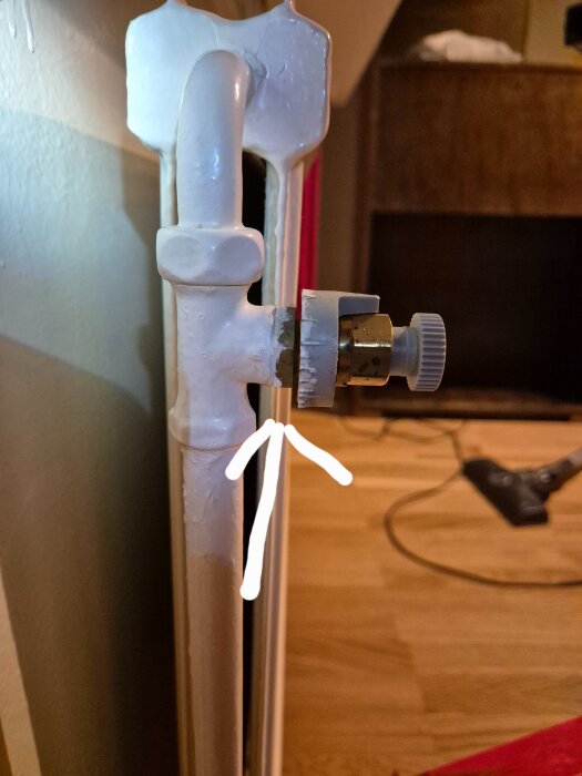 Vit målad radiator och rördel med ventil i fokus. En handritad pil pekar mot ventilen. Bakgrunden visar ett golv och oskarpa möbler.