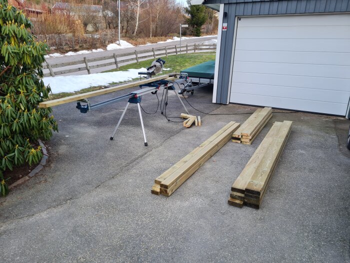 Tryckimpregnerade reglar staplade på en uppfart, med en kap- och gersåg på stativ i bakgrunden, redo för byggprojekt.
