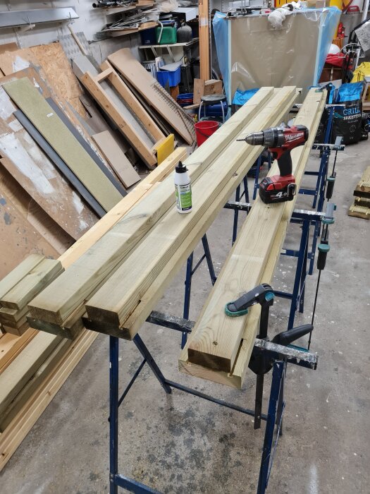 Tryckimpregnerade träreglar med urfrästa spår, en borrmaskin och en flaska lim på ett arbetsbord i en verkstad för byggprojekt.