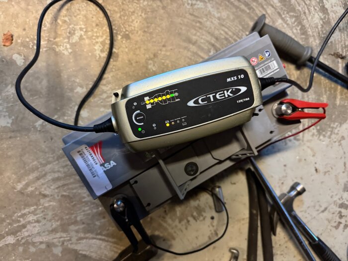 Batteriladdare av märket CTEK MXS 10 ansluten till ett bilbatteri med olika verktyg synliga runtom på ett betonggolv.