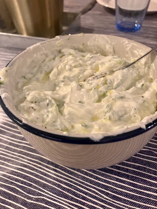 Skål med tzatziki på ett bord, krämig yoghurt med gurkbitar och kryddor synliga.