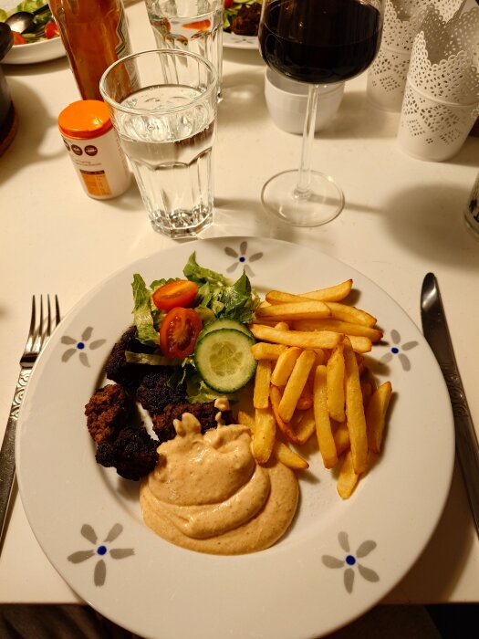 Kebab-biff med pommes frites, sås och grönsakssallad på en tallrik, med ett glas vin och vatten i bakgrunden.