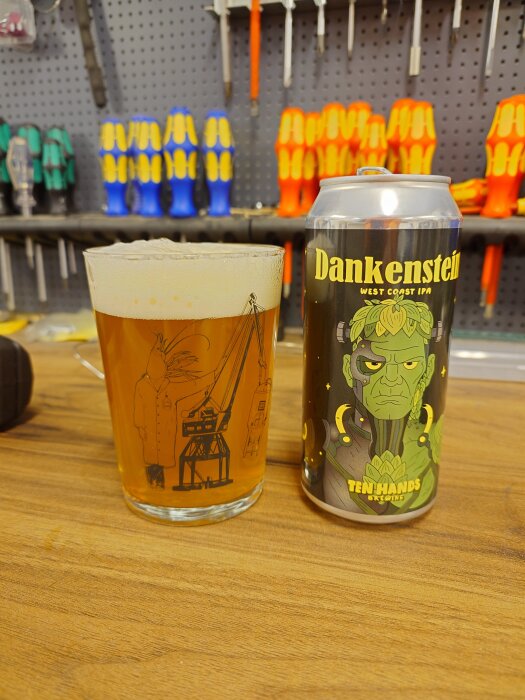 Ölglas med bärnstensfärgad dryck bredvid en burk märkt "Dankenstein West Coast IPA" på en arbetsbänk med verktyg i bakgrunden.