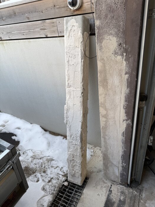 En riven ventilationstrumma i asbest står lutad mot en vägg, omgiven av snö och byggmaterial.