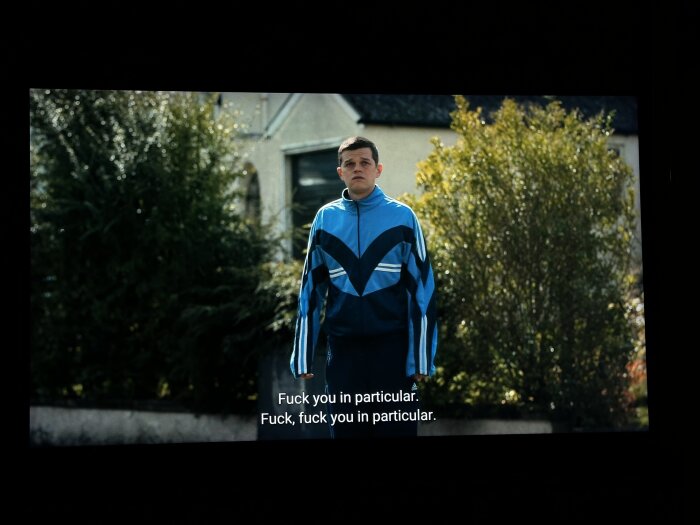 Ung man i blå träningsoverall står framför buskar, med textningen "Fuck you in particular" synlig.