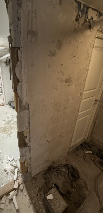Riven vägg med olika byggmaterial blottade, inklusive blåbetongblock och trä, omgiven av byggdamm och bråte på golvet.