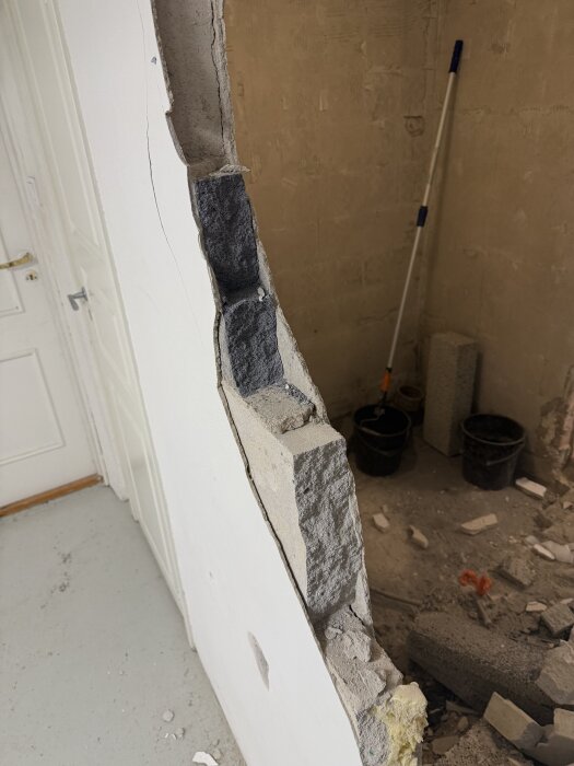 Riven vägg med synliga blåbetongblock och blandade material, verktyg och byggrester synliga i bakgrunden.
