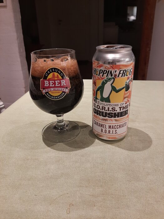 Ett glas mörkt öl bredvid en burk Hoppin' Frog B.O.R.I.S. The Crusher Caramel Macchiato står på ett bord med en grön duk.