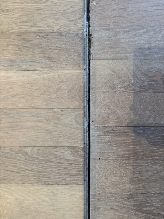 Golv med ett synligt glapp mellan två sektioner, den vänstra delen är i ljusare trä medan den högra är mörkare och har en skugga över sig.