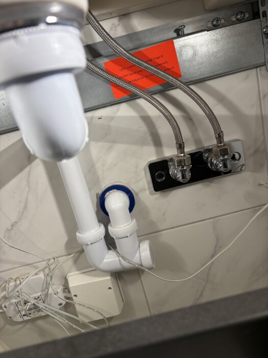 Kopplingar och avloppsrör under diskbänk för inkoppling av tvättmaskinens kallvattenslang, med två metallslangar och el i bakgrunden.