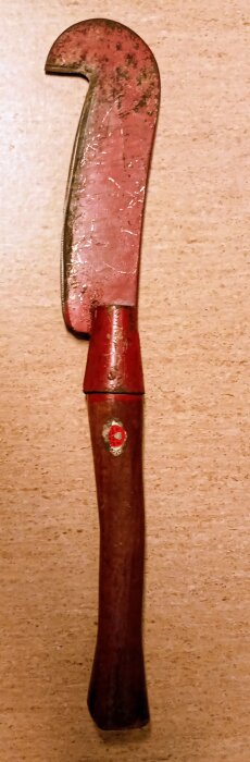 Gammal rödmålad trähacka eller dräktkniv med trähandtag, liggande på en ljus yta. Verktyget är slitet och har en krokig klinga.