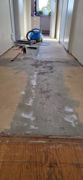 En hall med borttagen plastmatta, synliga spacklade skarvar och verktyg för golvrenovering. En dammsugare står i bakgrunden.