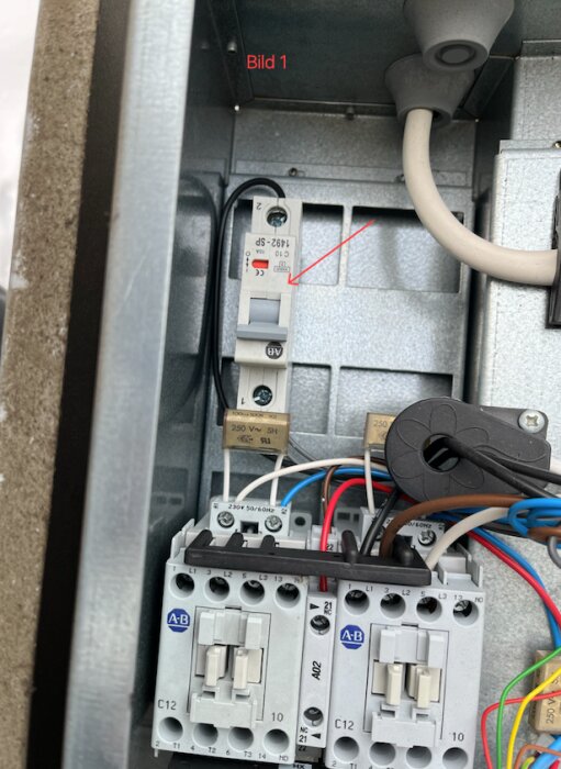 Säkring på tryckets högra sida i ett elskåp, markerad med röd pil. Den står i "1" läge med röd indikator synlig, omgiven av kablar och komponenter.