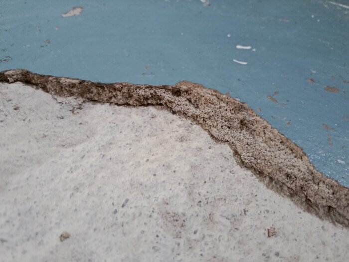 Betonggolv med avlägsnat ytskikt, där ett område på 1-2,5 cm saknas, exponeras. Underlag delvis blåmålat. Från en källarrenovering.