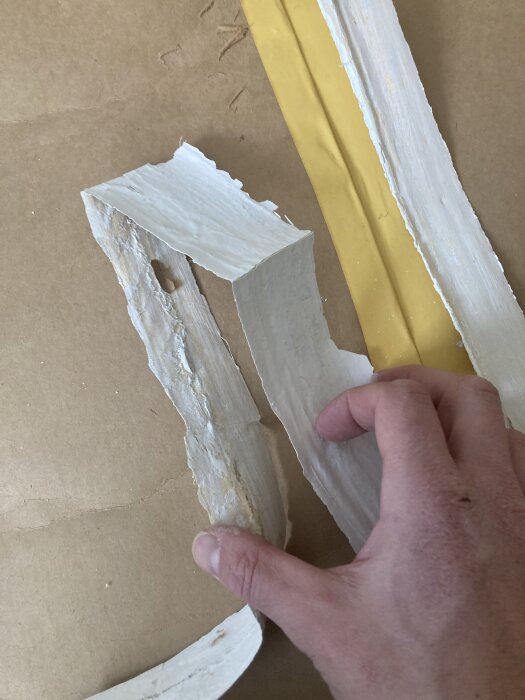 Hand som håller i flagnad färg och papper från ett gammalt spänntak med vattenskada, på en brun yta.