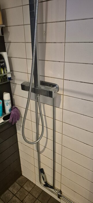 Duscharmatur som är monterad på kaklad vägg med en flexibel duschslang och takdusch.
