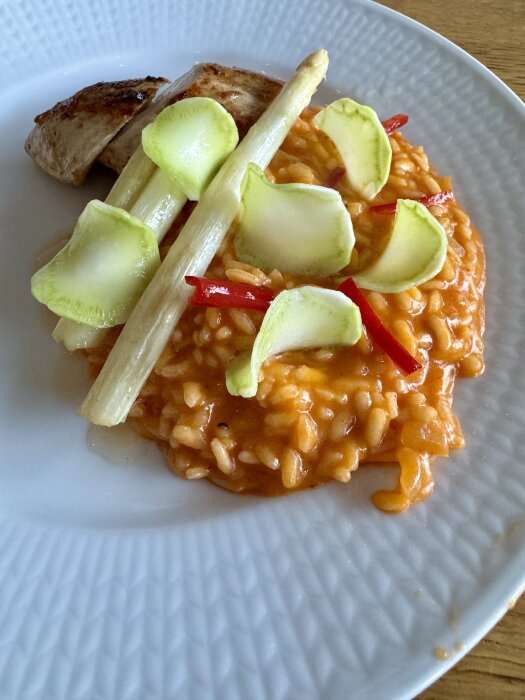 Tomatrisotto med kyckling, vit sparris, syrad broccolistam, chili och parmaskinka serverad på en vit tallrik.