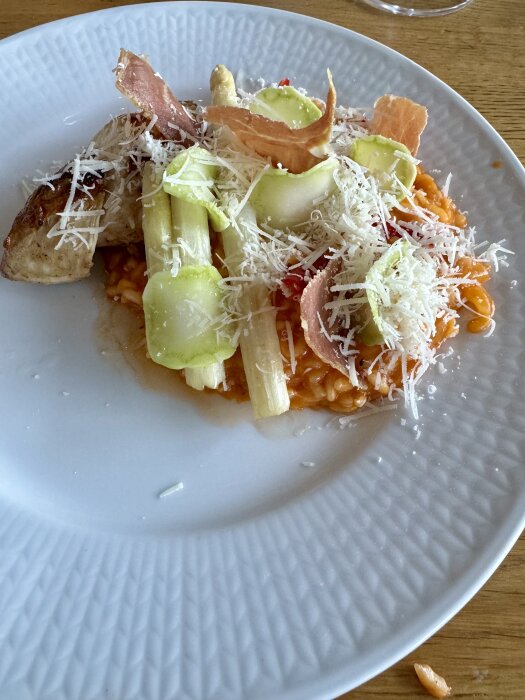 Risotto toppad med sparris, skivad squash, lufttorkad skinka och riven parmesan på en vit tallrik.