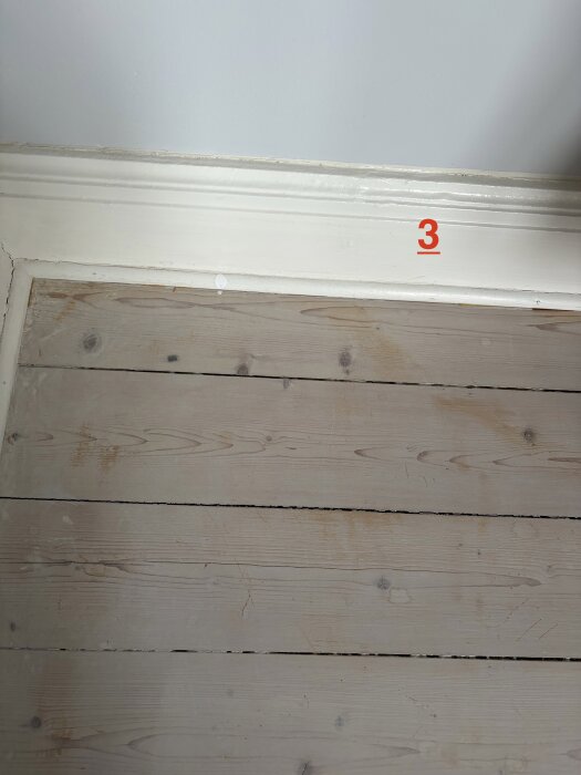 Slitet golv av träplankor, ljus färg med sprickor och smutsfläckar, nära vägglist, markerad med röd siffra 3 på golvet.