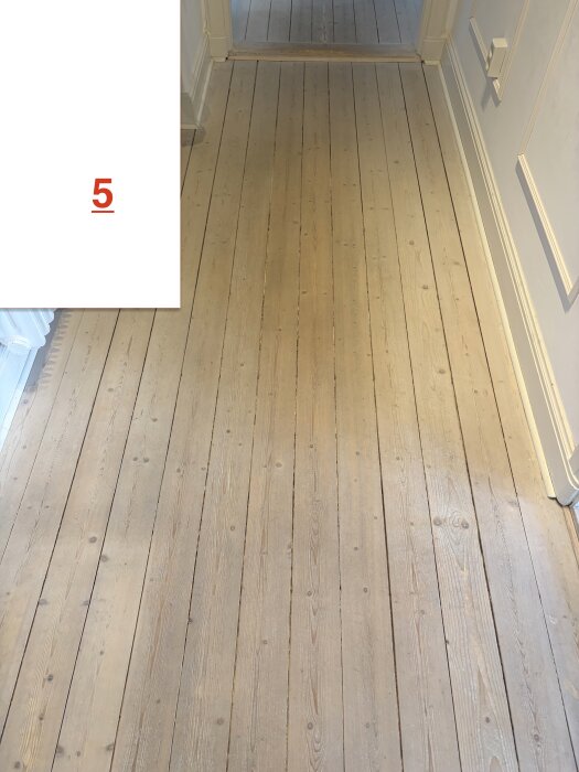 Ljusa trägolvplankor i en hall, med vita väggar och dörrkarmar.