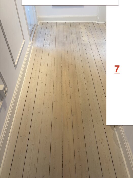 Ljust trägolv i en hall med vita väggar och dörrkarmar.