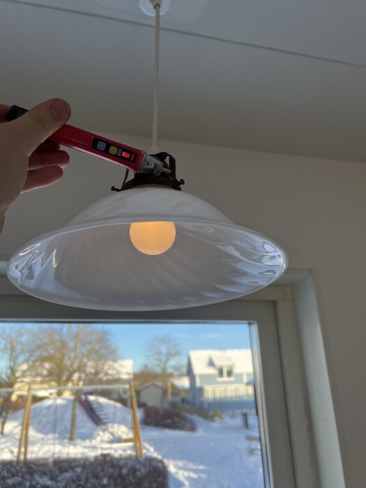 Glödande dimbar LED-lampa i en taklampa, en provpenna hålls nära lampans sockel i ett kök med fönster utsikt över snötäckt trädgård.