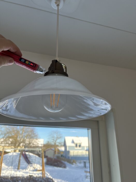 Hand använder en provpenna på en taklampa med dimbar LED-lampa i ett kök, med snötäckt utsikt i bakgrunden.