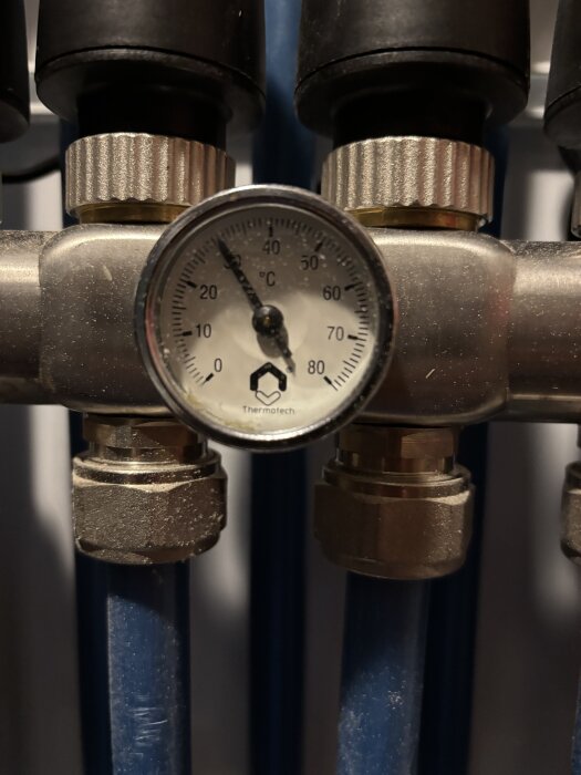 Termometer på rörledning för vattenburen golvvärme, visar cirka 20°C, omgiven av metallkopplingar och blå plastslangar.