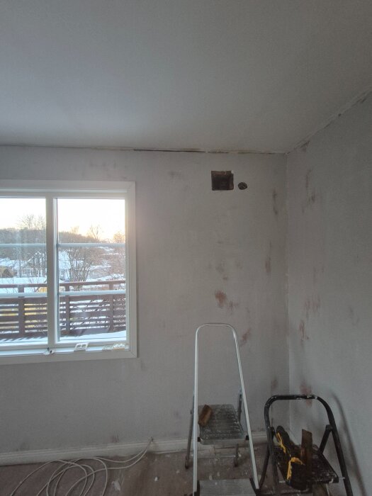 Öppning i väggen nära taket i ett rum med stege och renoveringsverktyg, bredvid ett fönster med utsikt över snötäckt landskap.