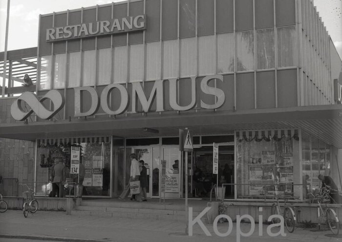 Svartvit bild av Domus Timrå med restaurangskylt ovanför ingången. Flera personer rör sig framför butiken. Cyklister och fönster med affischer syns.