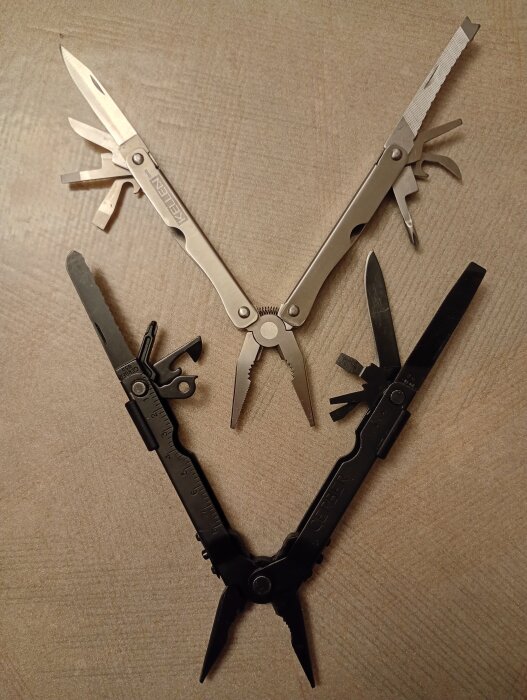Två multiverktyg med olika verktyg utfällda, ett silverfärgat märkt "Kellen" och ett svart. Verktygen ligger på ett träbord.