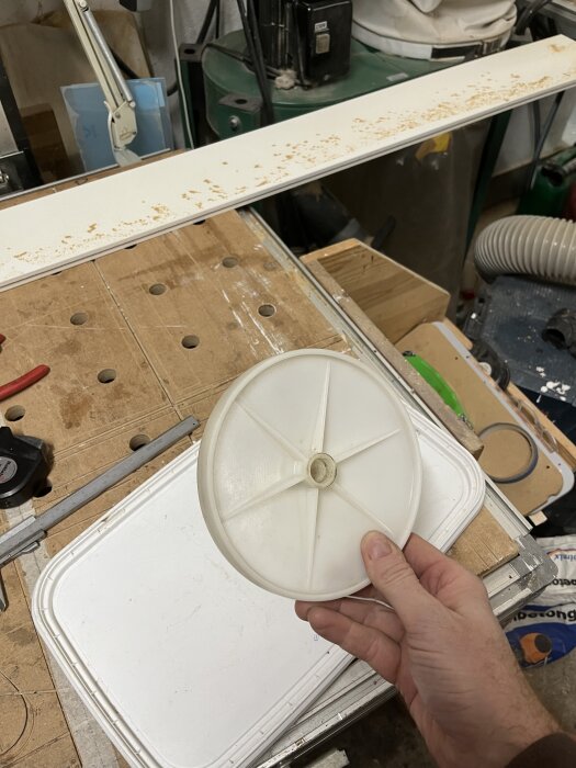 En person håller ett vitt plastlock i en verkstad med verktyg och material i bakgrunden.