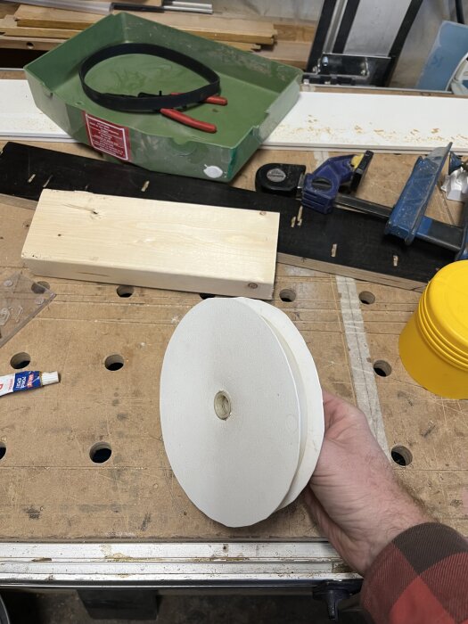 Hand som håller ihoplimmade lock från målarhinkar; verktyg och material ligger på arbetsbänk i bakgrunden.