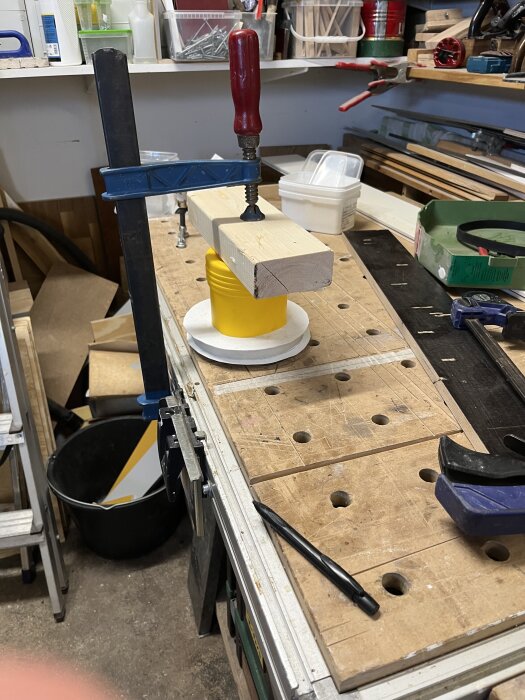 Arbetsbänk med ett DIY-projekt där två lock från målarhinkar är fastklämda med en träbit och skruvtving i en arbetsstation.