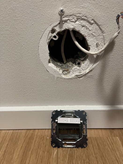 Öppet vägguttag med lösa kablar och ett olöst RJ45-uttag som ligger på golvet framför hålet i väggen.