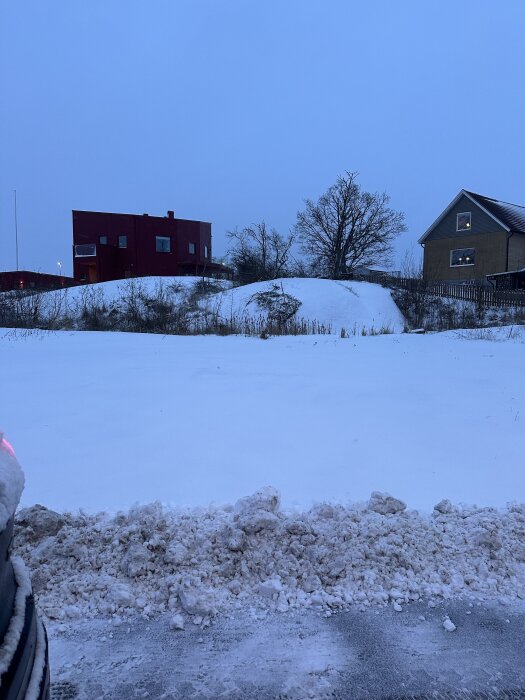 Snötäckt mark med stugor i bakgrunden, snövall i förgrunden, och träd.