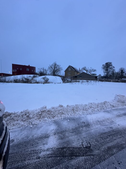 Snötäckt tomt med utsikt mot ett berg i bakgrunden samt ett gult hus och träd under en molnig himmel.
