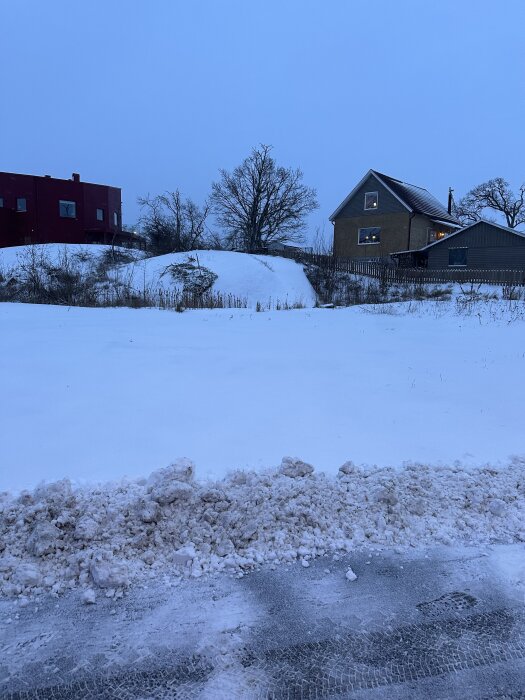 Snötäckta tomter med två hus i bakgrunden och ett lågt berg på högra sidan, omgivna av några träd.