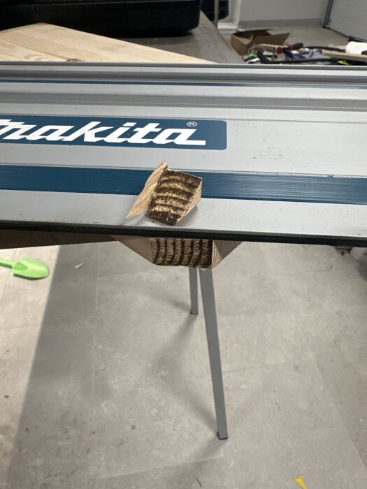 Sågspår på en träbit placerad mot en Makita sågbänkskant, med verktyg i bakgrunden på ett grått golv.