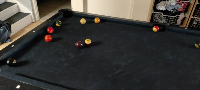 Biljardbord med svart filt och färgglada bollar, nära en trappa och korgar med kläder i bakgrunden.