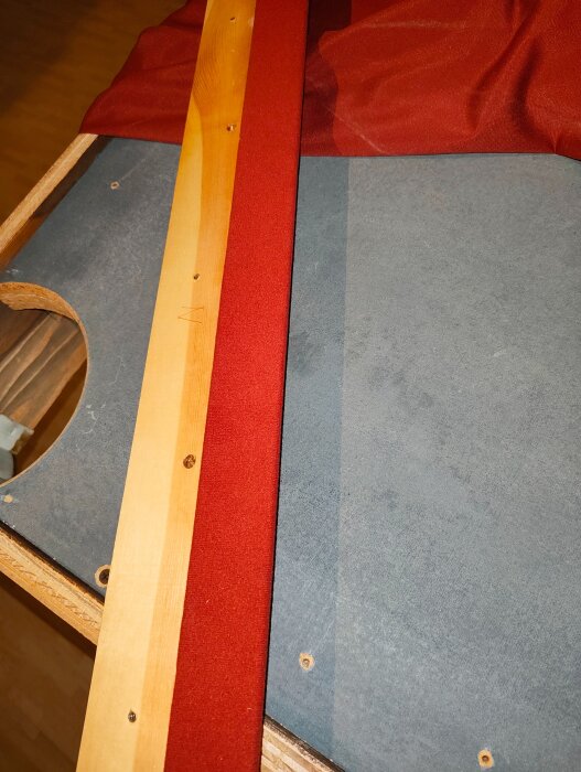 En närbild av ett biljardbord under renovering med en röd duk som delvis täcker bordets yta och trädetaljer synliga.