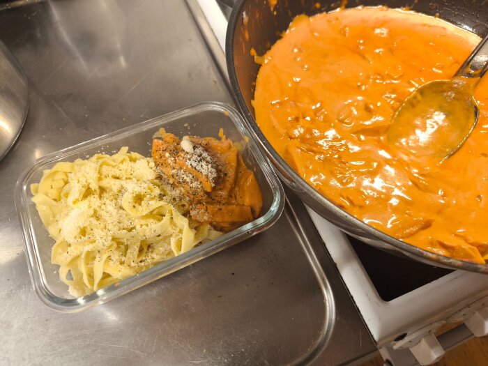 Matlåda med korvstroganoff och färsk pasta toppad med parmesan, bredvid en stekpanna med mer korvstroganoff på en spis.