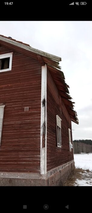 Rött trähus med skadat tak och vägg, beläget i en snötäckt landskap. Träpanelen är sliten och färgen flagnad.