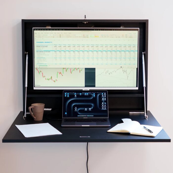 Laptop på skrivbord med anteckningar och penna, uppfällbar skärm med grafer och data ovanför. Kopp vid sidan om datorn.