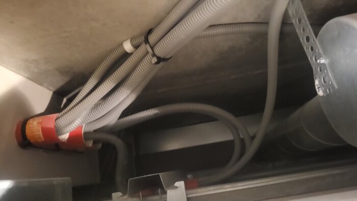 Bilden visar flera gråa, korrugerade slangar som är sammanknutna och passerar genom en vägg tillsammans med ett rött rör.