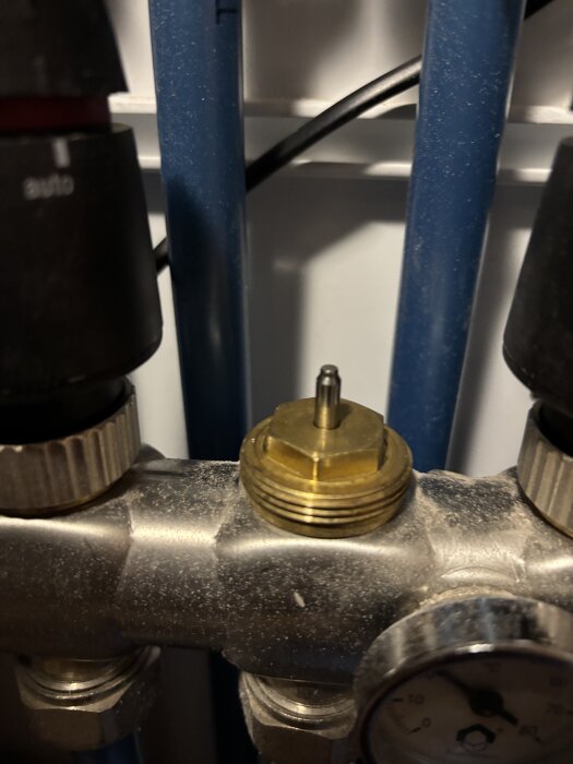 Närbild på VVS-installation med mässingskomponent och manometer omgiven av blå rör och svarta vridhandtag.