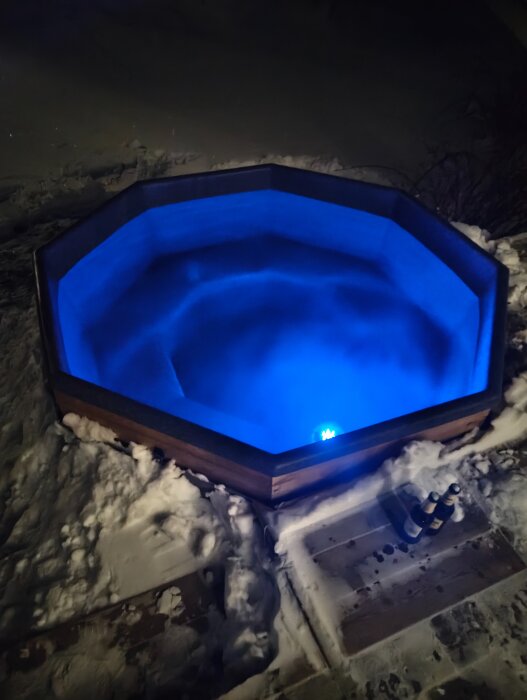 Rund badtunna med blå belysning utomhus i snön, två flaskor vid kanten.